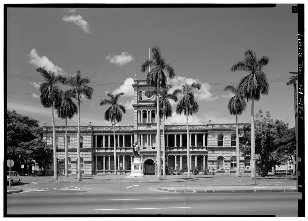 Aliiolani Hale behind palm trees, with a central clock tower and the statue of King Kamehameha I in front.