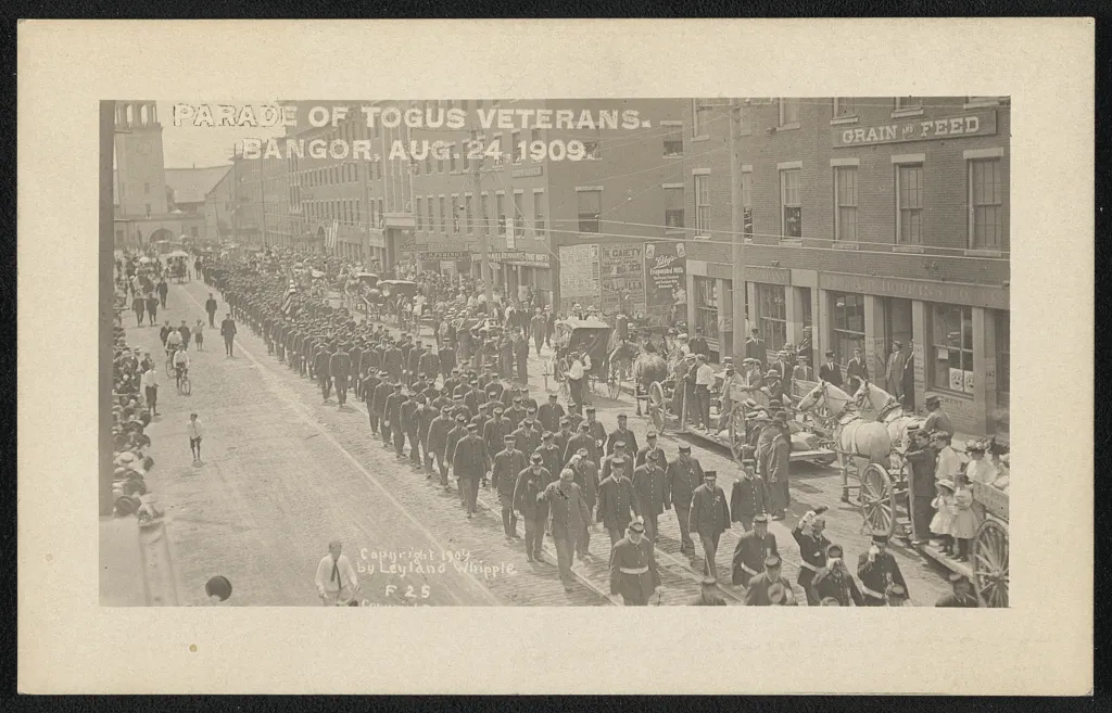 A 1909 Bangor street lined with spectators as uniformed veterans march past commercial buildings.