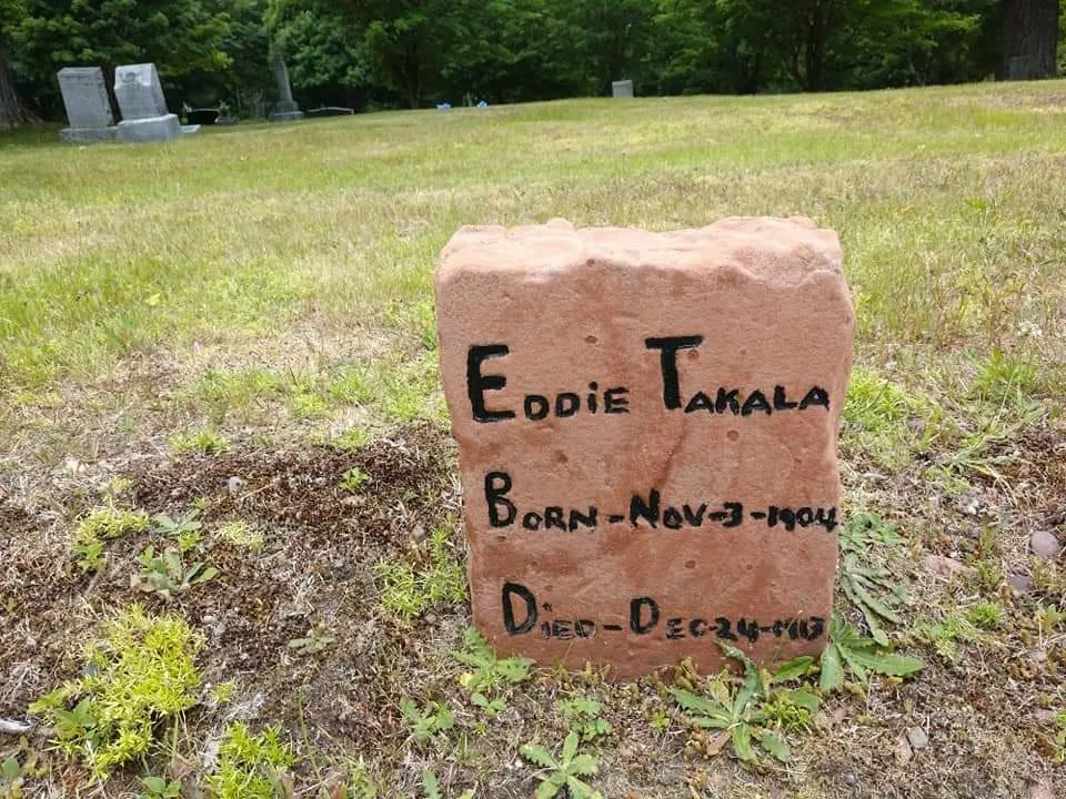 Eddie Takala's handmade sandstone grave marker on a grassy cemetery slope, with his name and 1904 and 1913 dates.