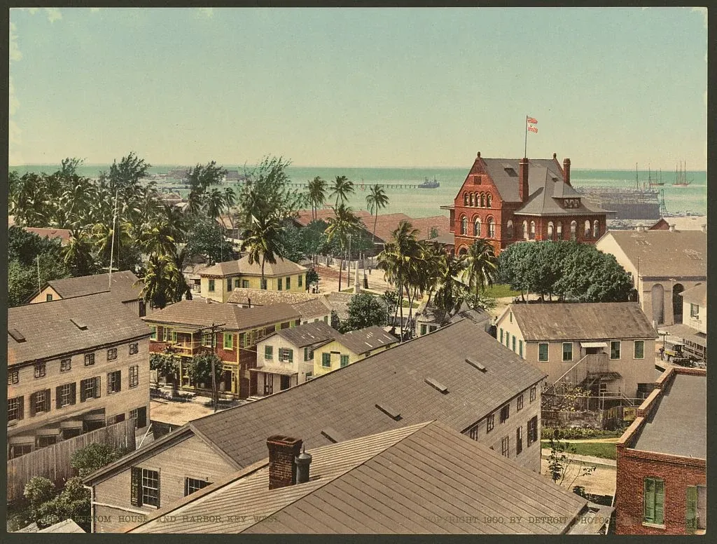 Historic color print of Key West rooftops, palm trees, the red brick Custom House and ships in the harbor.