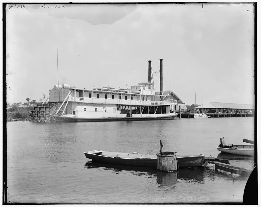 Black and white photograph of a large river steamboat with twin smokestacks, small rowboats and waterfront docks in Miami.