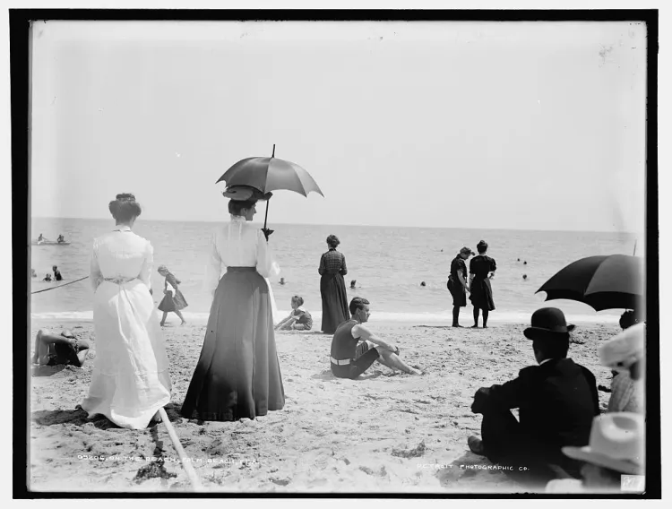 Black and white photograph of visitors in early twentieth century clothing standing and sitting on Palm Beach, with parasols and bathers in the sea.