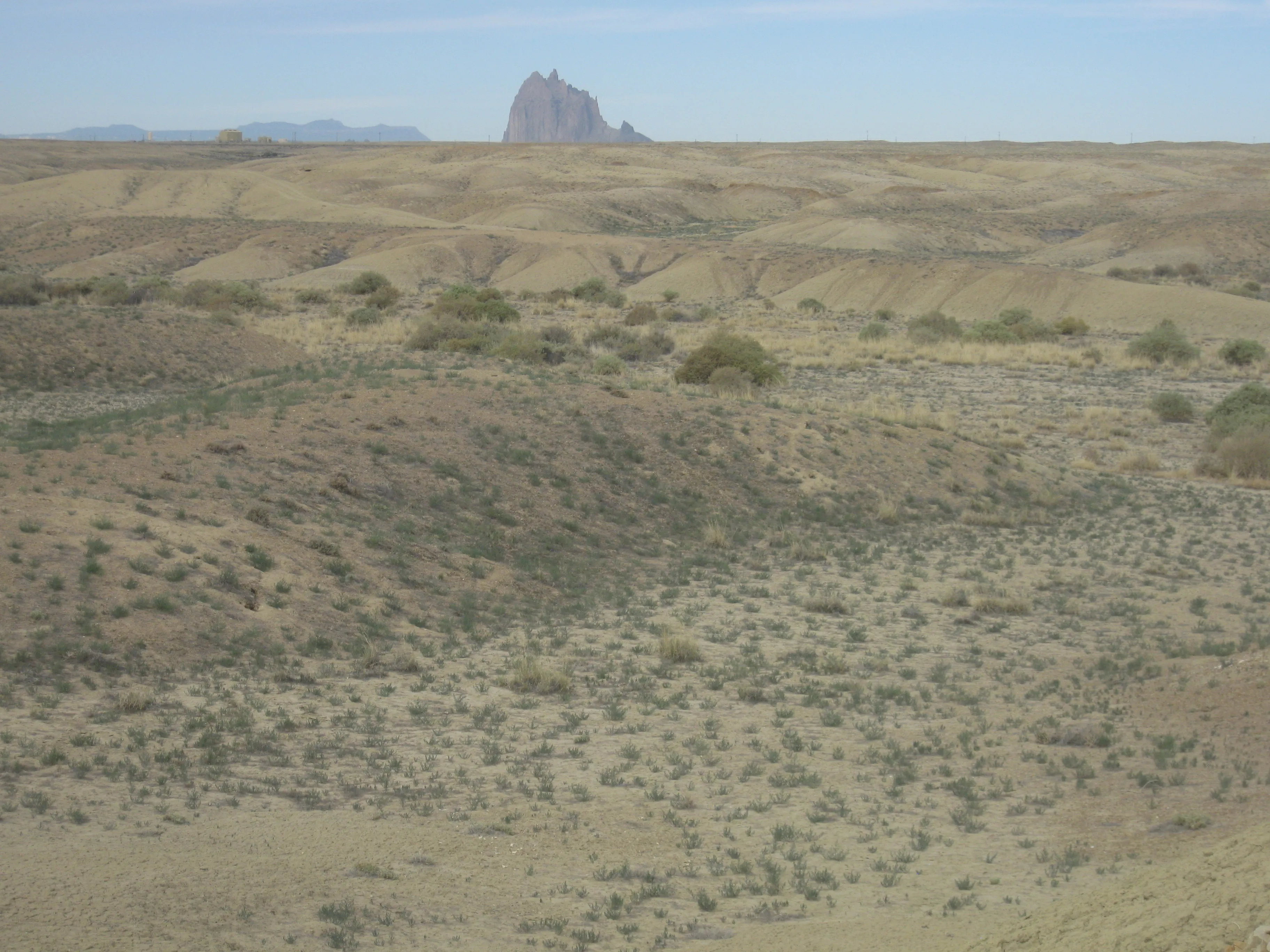 Dry, ridged terrain with Shiprock rising in the distance beneath a pale sky